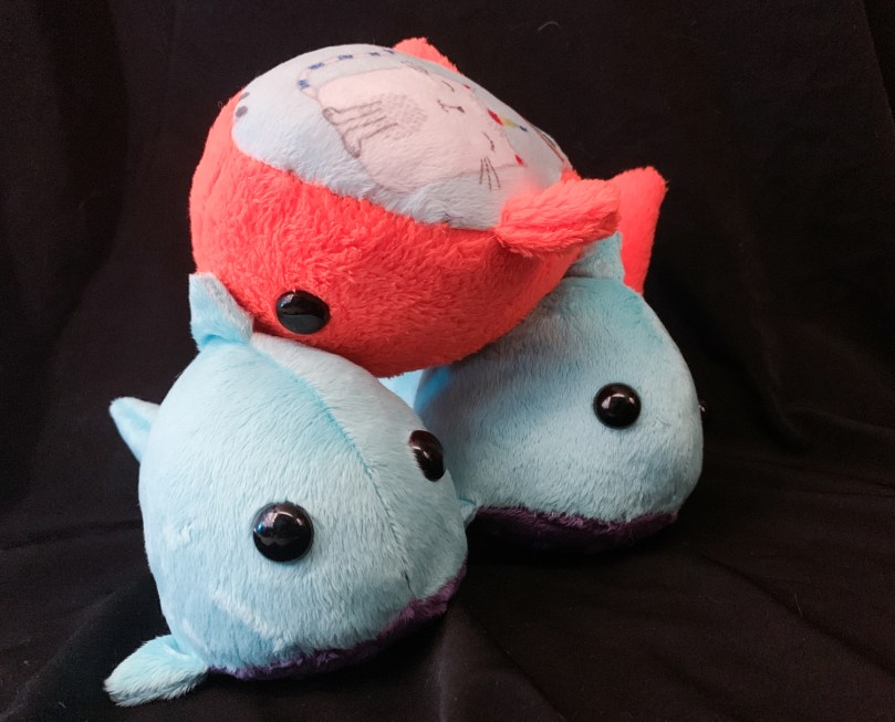 three stuffed sharks, two are blue with purple bellies and one is orange with a cat on its belly. The orange one is upside down on top of the other two, which are right side up.